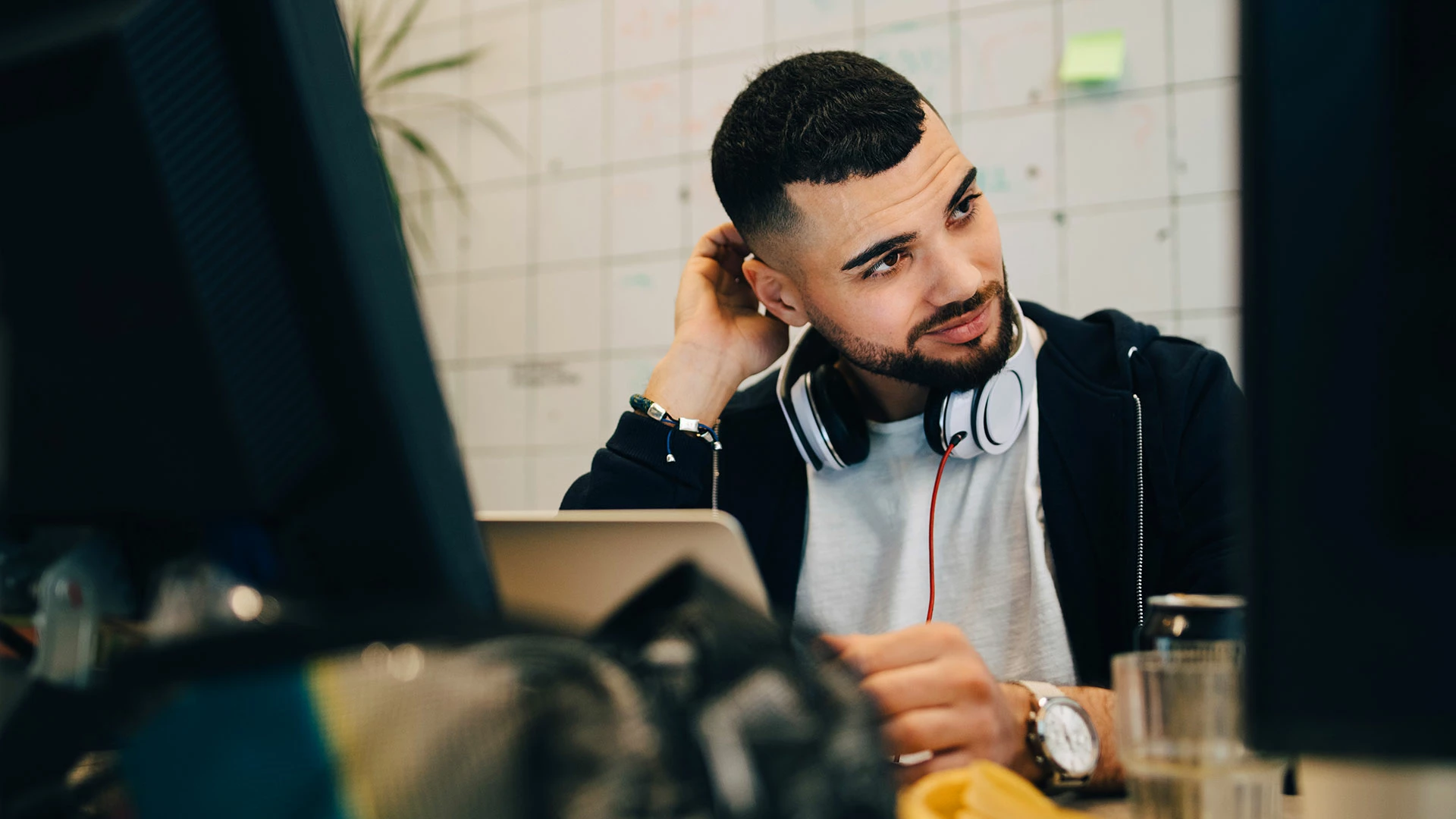 Confused young male computer programmer scratching head while sitting with laptop at creative office 